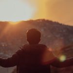 A man stands with open arms, silhouetted against a vibrant sunset over a mountainous landscape.
