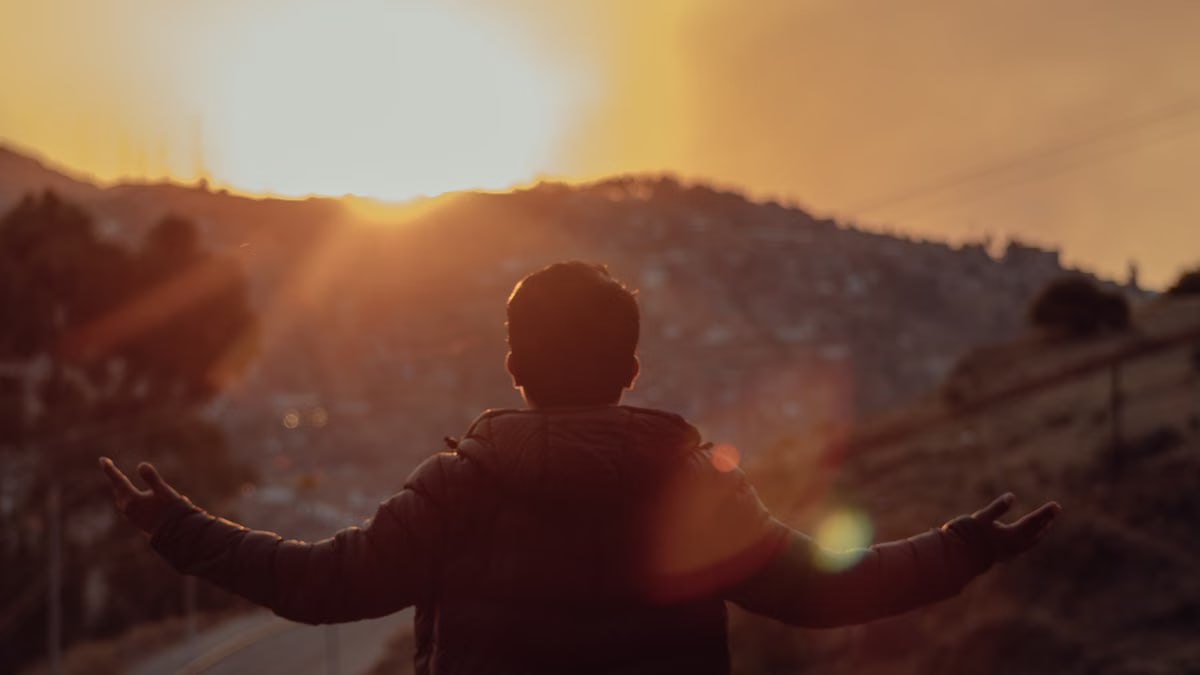 A man stands with open arms, silhouetted against a vibrant sunset over a mountainous landscape.